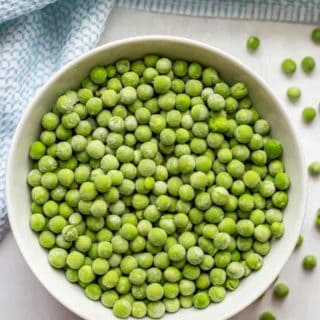 Frozen green peas in a white bowl with extra peas scattered nearby