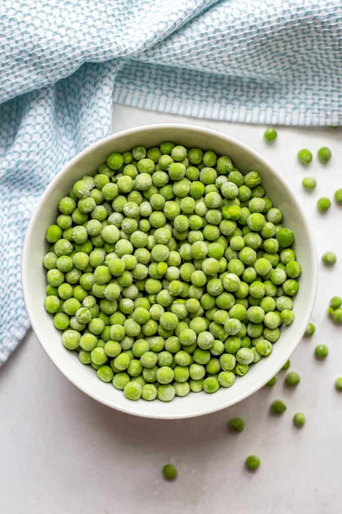 A white bowl full of frozen green peas with peas scattered around on the counter