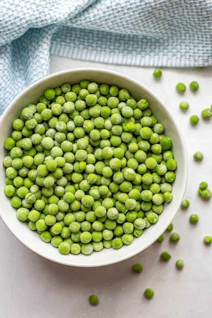 A white bowl full of frozen green peas with peas scattered around on the counter