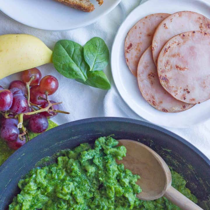 Scrambled green eggs in a saute pan with toast, ham and fruit nearby