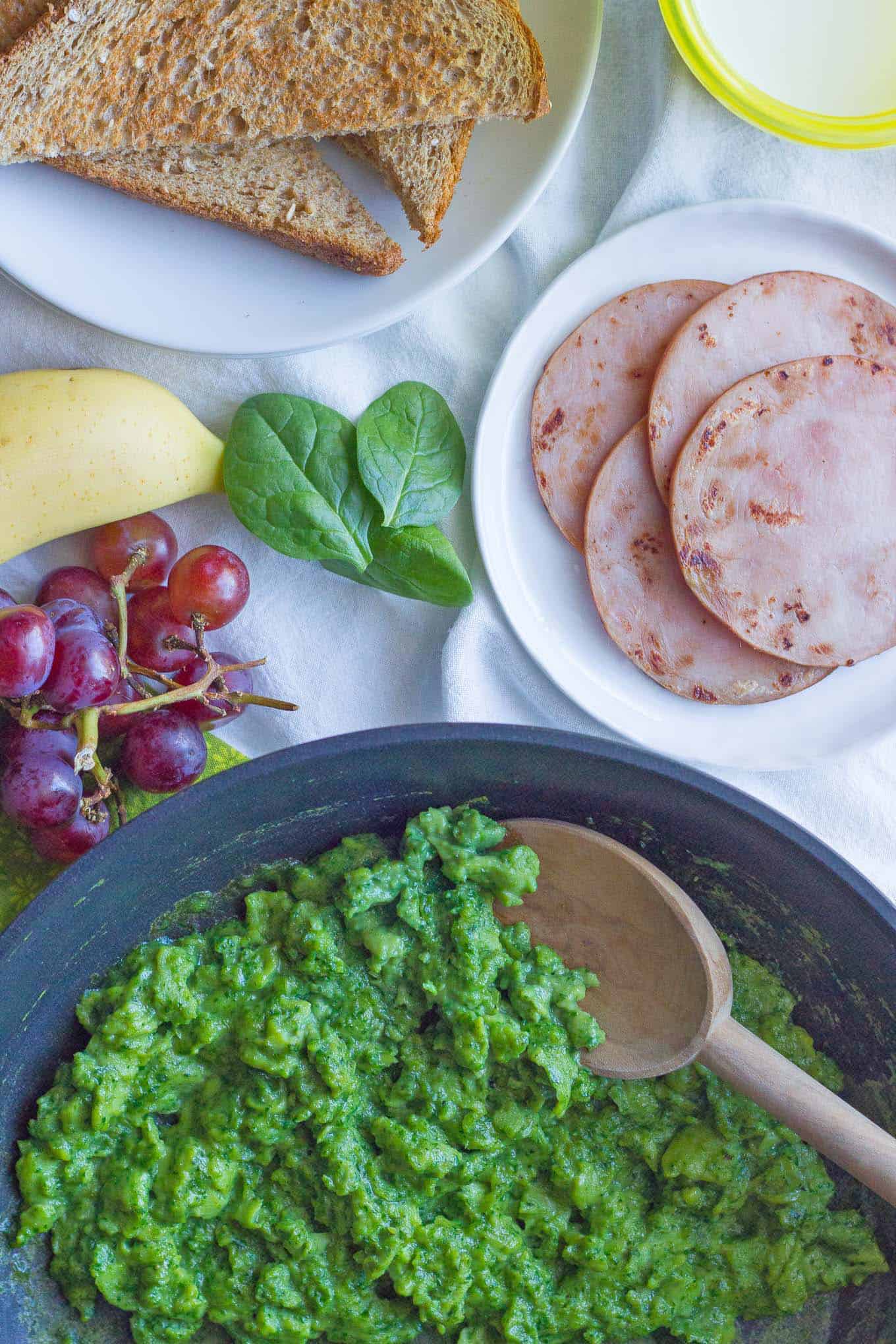 Scrambled green eggs in a saute pan with toast, ham and fruit nearby