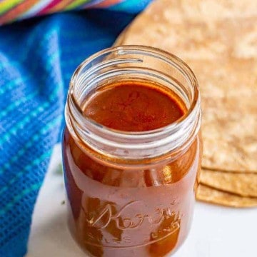 A glass jar filled with homemade enchilada sauce with tortillas and cilantro on the counter nearby