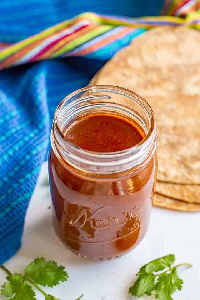 A glass jar filled with homemade enchilada sauce with tortillas and cilantro on the counter nearby