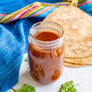 A glass jar with homemade enchilada sauce with some stacked tortillas and a colorful blue towel nearby