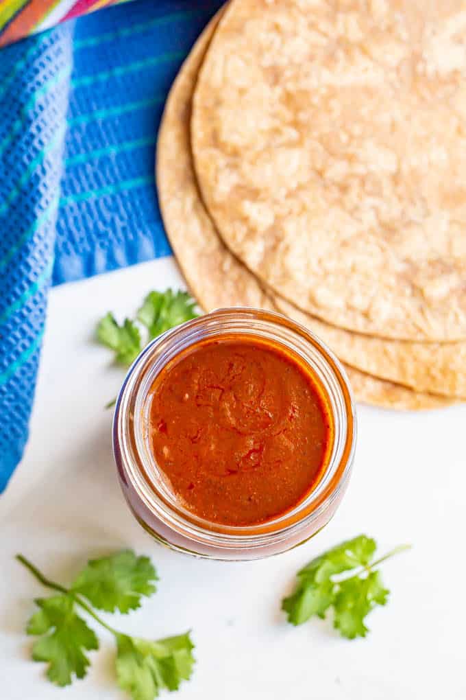 Overhead image of red enchilada sauce in a glass jar with some tortillas stacked nearby