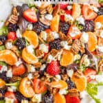 Close up of a mixed greens salad with strawberries, blackberries and mandarin oranges plus nuts and crumbled goat cheese in a large white oval serving platter.