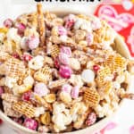 A white chocolate coated Valentine's themed chex mix with colored m&ms and pretzels in a large white bowl.