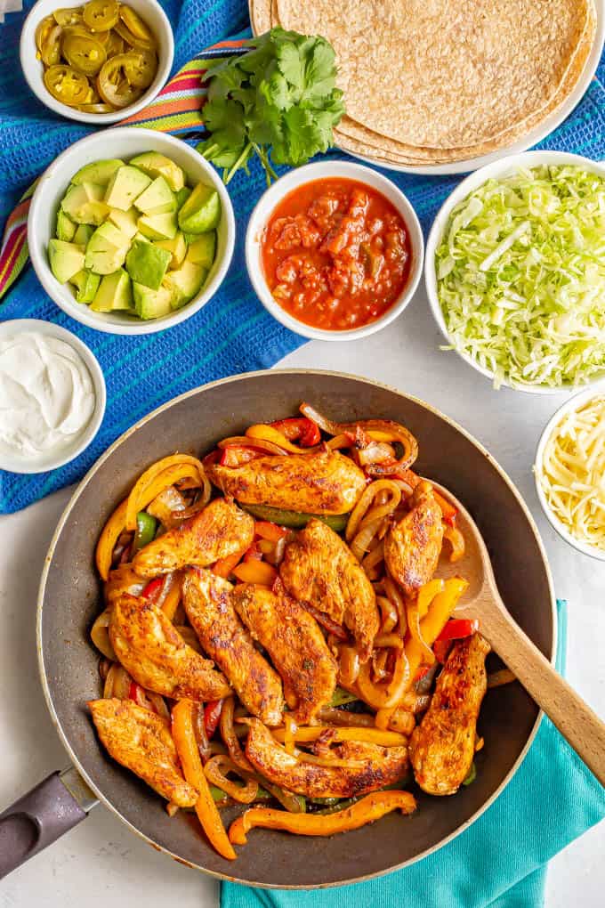 A skillet of fajita chicken and peppers surrounded by bowls of toppings and a plate of flour tortillas