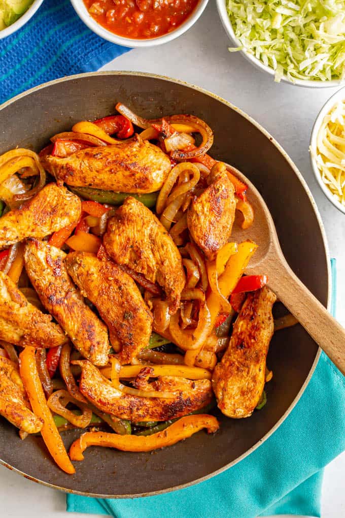 A wooden spoon scooping up some fajita chicken and peppers from a skillet, with bowls of toppings in the background