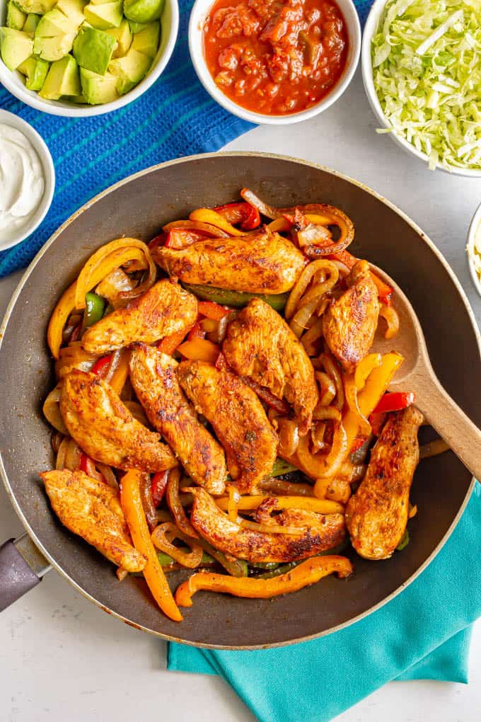 A skillet of cooked chicken fajitas with bell peppers and onions and bowls of toppings nearby