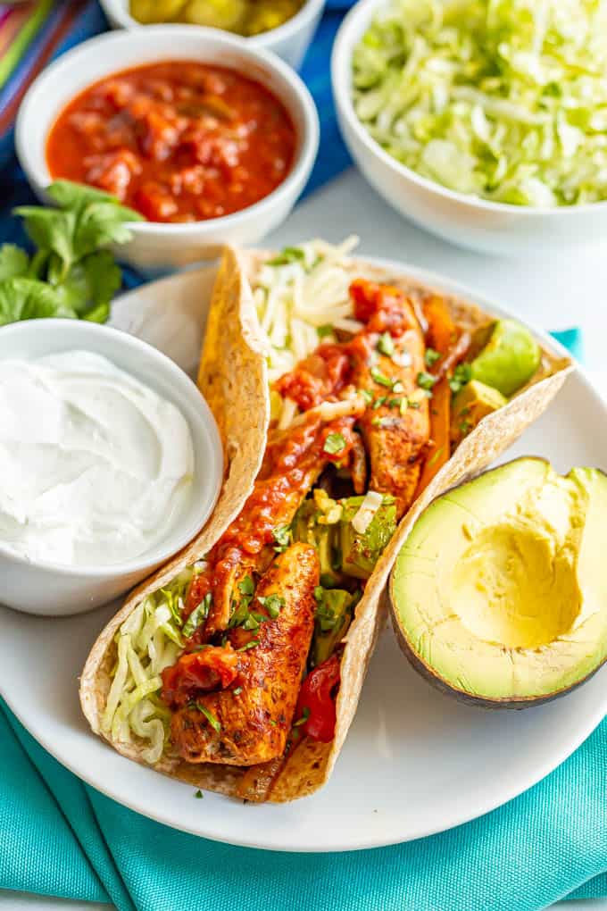 A loaded chicken fajita wrapped in a tortilla and served on a white plate with avocado, salsa and toppings nearby