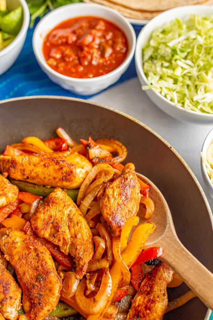 A wooden spoon scooping up some fajita chicken and peppers from a skillet, with bowls of toppings in the background