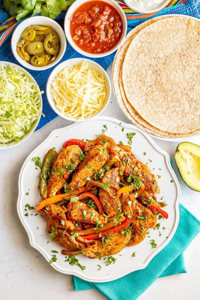 A large white plate piled high with chicken fajitas with small bowls of toppings and a plate of flour tortillas nearby