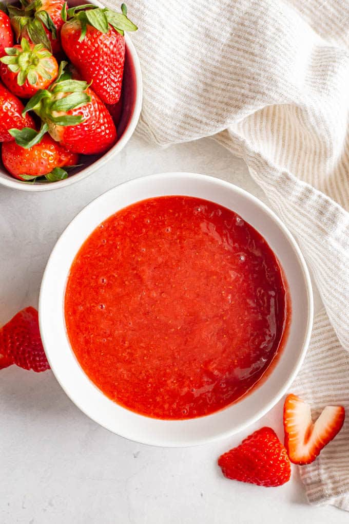 A white bowl with homemade strawberry sauce and a bowlful of strawberries nearby