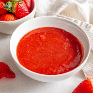A white bowl with bright strawberry sauce and a bowlful of strawberries nearby