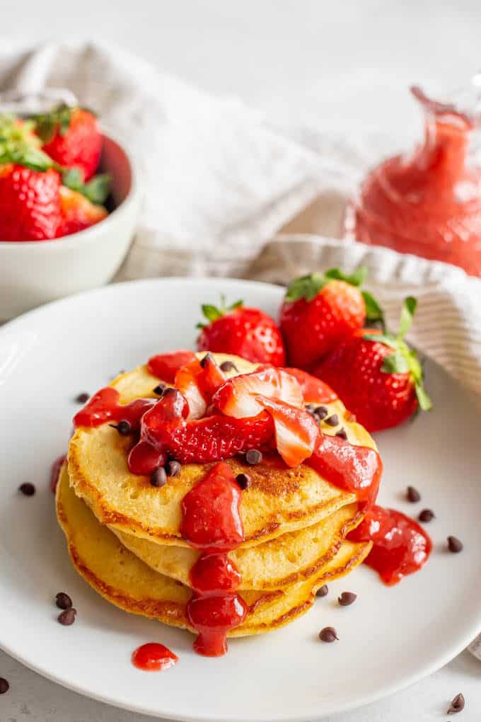 Pancakes stacked on a white plate topped with mini chocolate chips and strawberry sauce and a bowl of strawberries in the background