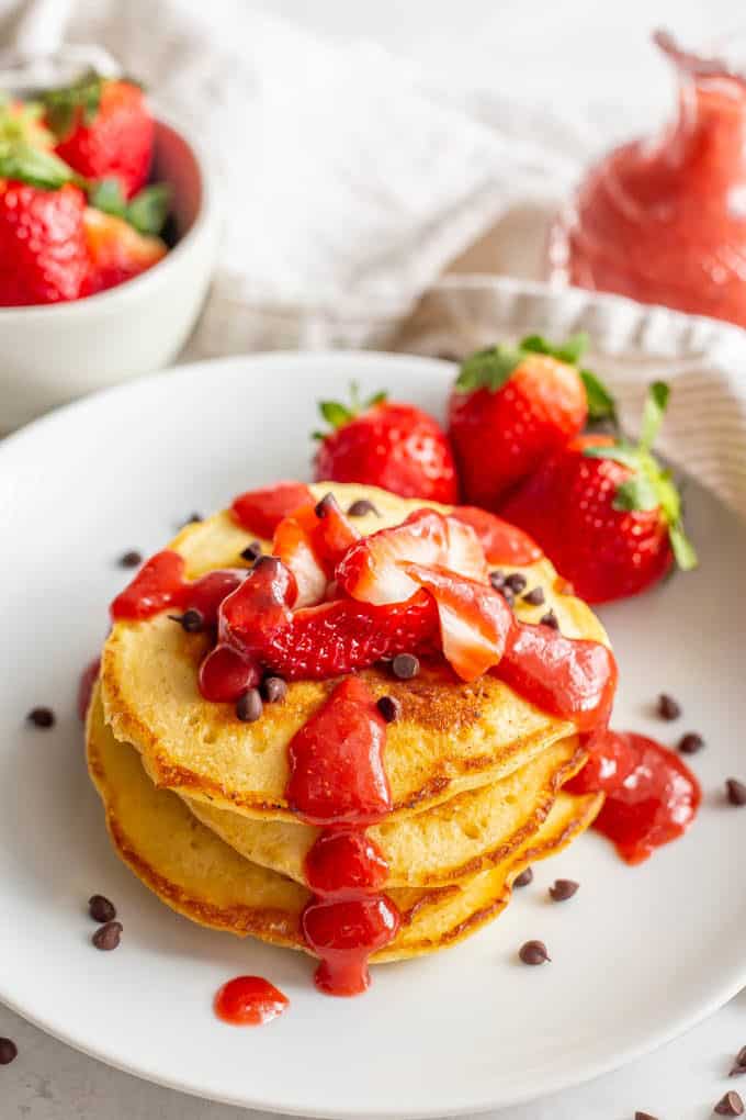 Pancakes stacked on a white plate topped with mini chocolate chips and strawberry sauce