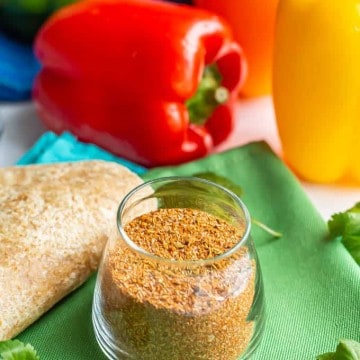 Homemade fajita seasoning in a small glass jar with flour tortillas, bell peppers and cilantro sprigs nearby