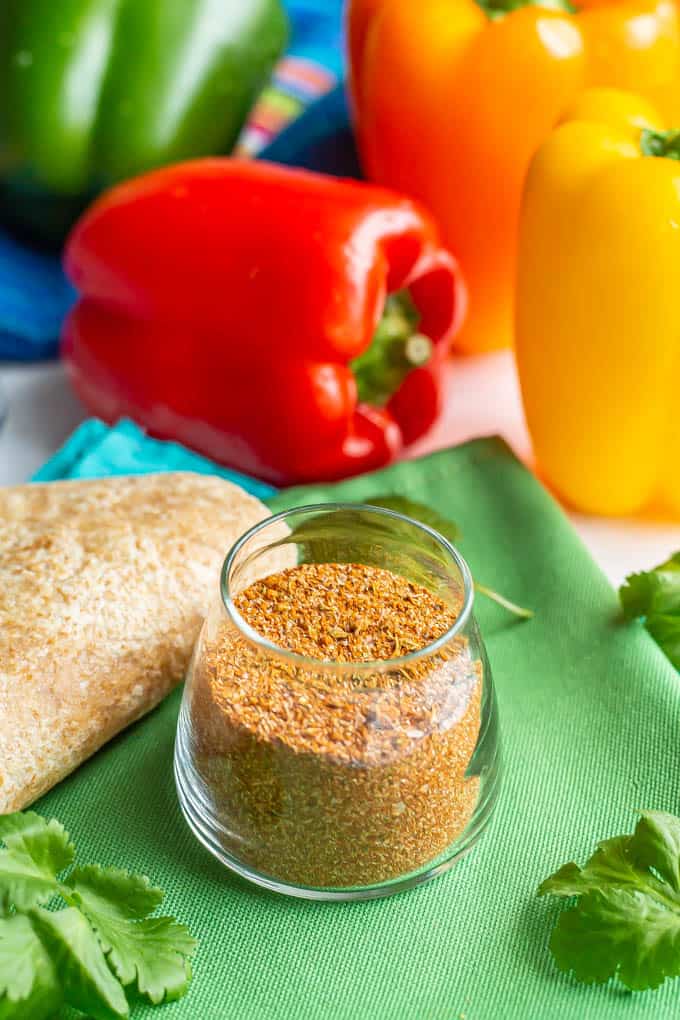 Homemade fajita seasoning in a small glass jar with flour tortillas, bell peppers and cilantro sprigs nearby