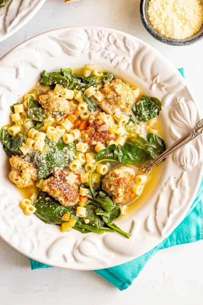 A decorative white bowl full of soup with meatballs, pasta and spinach and a spoon resting on the side