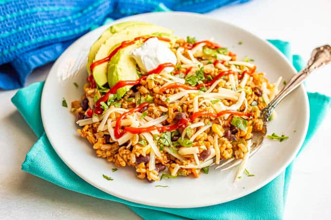 Mexican fried rice served on a white plate with a fork and topped with cilantro, shredded cheese, avocado and sriracha sauce