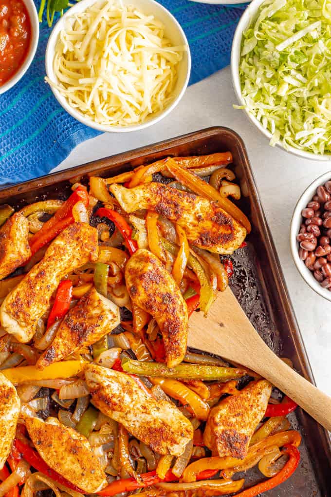 A wooden spatula scooping up chicken fajitas with bell peppers and onions from a baking sheet and nearby bowls of toppings