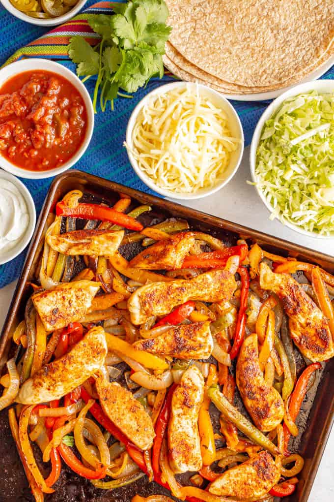 Baked chicken fajitas with bell peppers and onions and nearby bowls of toppings and a plate of tortillas