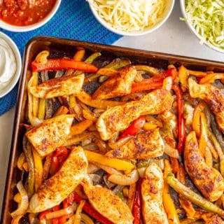 Sheet pan chicken fajitas with bell peppers and onions and nearby bowls of toppings
