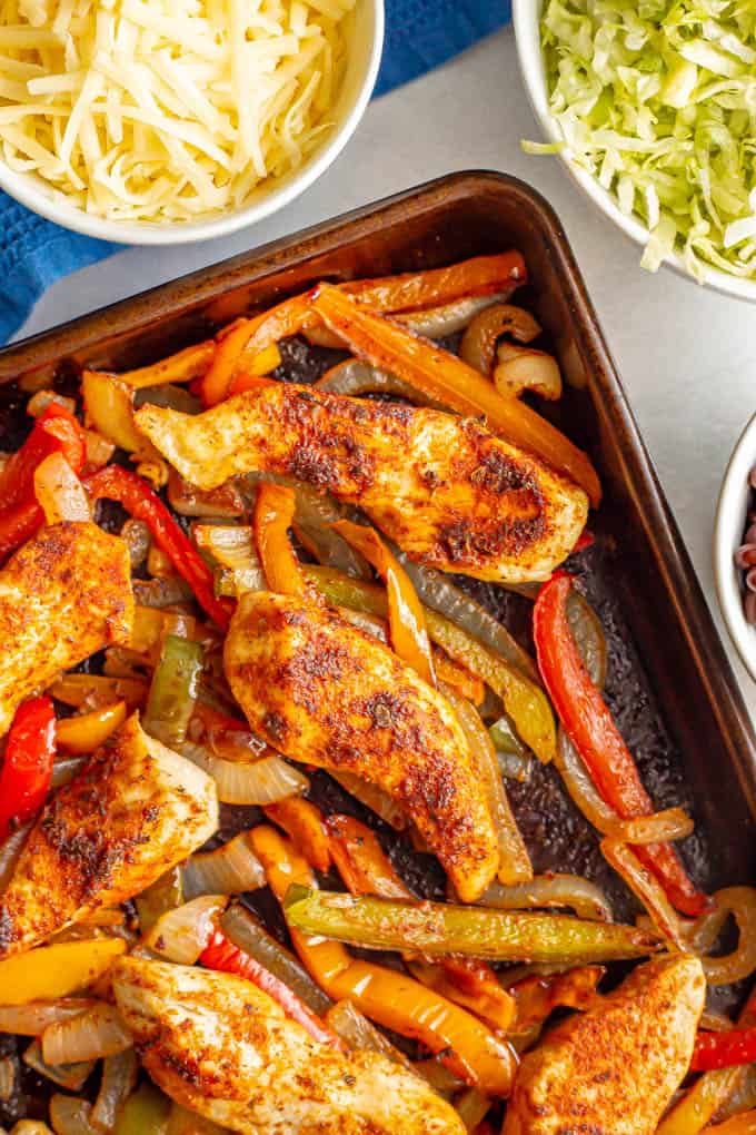 Sheet pan chicken fajitas with bell peppers and onions and nearby bowls of toppings