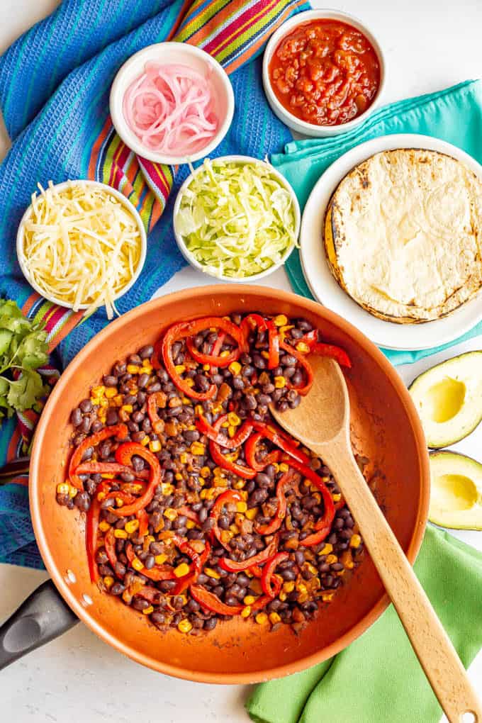 A skillet with black beans, peppers and corn surrounding by taco toppings in separate bowls