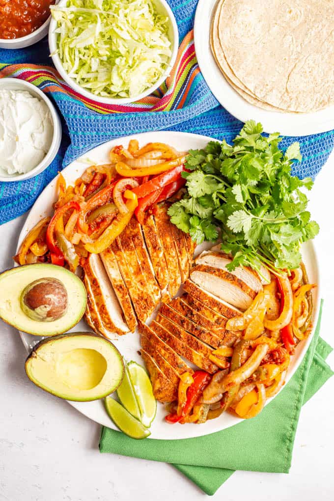 A large white serving platter piled with crock pot chicken fajitas plus garnishes and bowls of toppings nearby