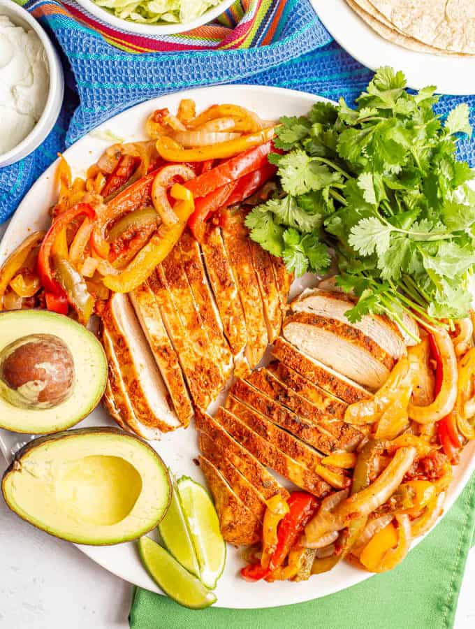A large white serving platter piled with fajita chicken and peppers and onions, plus garnishes and bowls of toppings nearby
