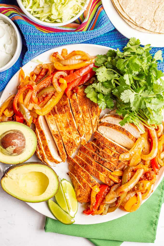 A large white serving platter piled with fajita chicken and peppers and onions, plus garnishes and bowls of toppings nearby