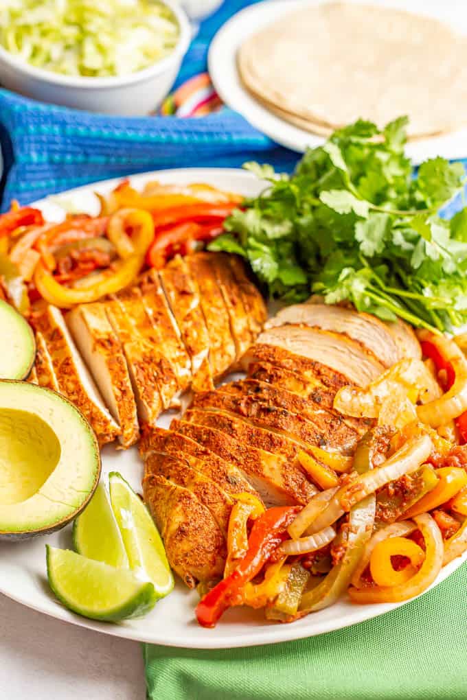 A large white serving platter piled with fajita chicken and peppers and onions, plus lime, avocado and cilantro