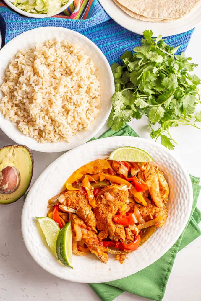 Fajita chicken and bell peppers in a white serving bowl with lime wedges and a bowl of rice and some cilantro in the background