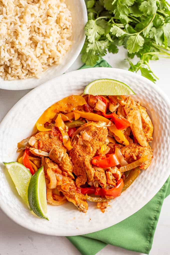 Chicken and bell pepper fajitas in a white serving bowl with lime wedges and a bowl of rice and some cilantro in the background