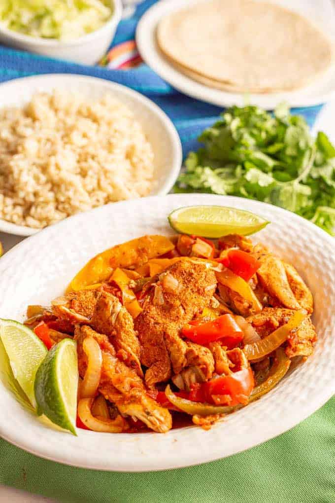 Chicken and bell pepper fajitas in a white serving bowl with lime wedges and a bowl of rice in the background