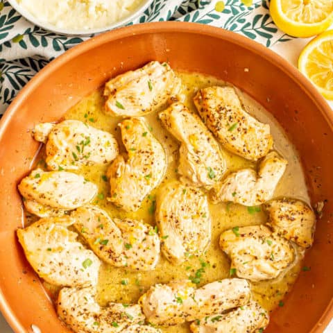 Cooked chicken strips in a lemon butter sauce in a copper skillet with a bowl of mashed potatoes in the background