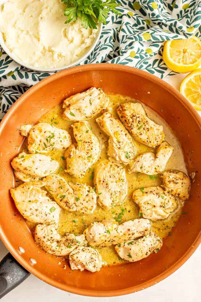 Cooked chicken strips in a lemon butter sauce in a copper skillet with a bowl of mashed potatoes in the background