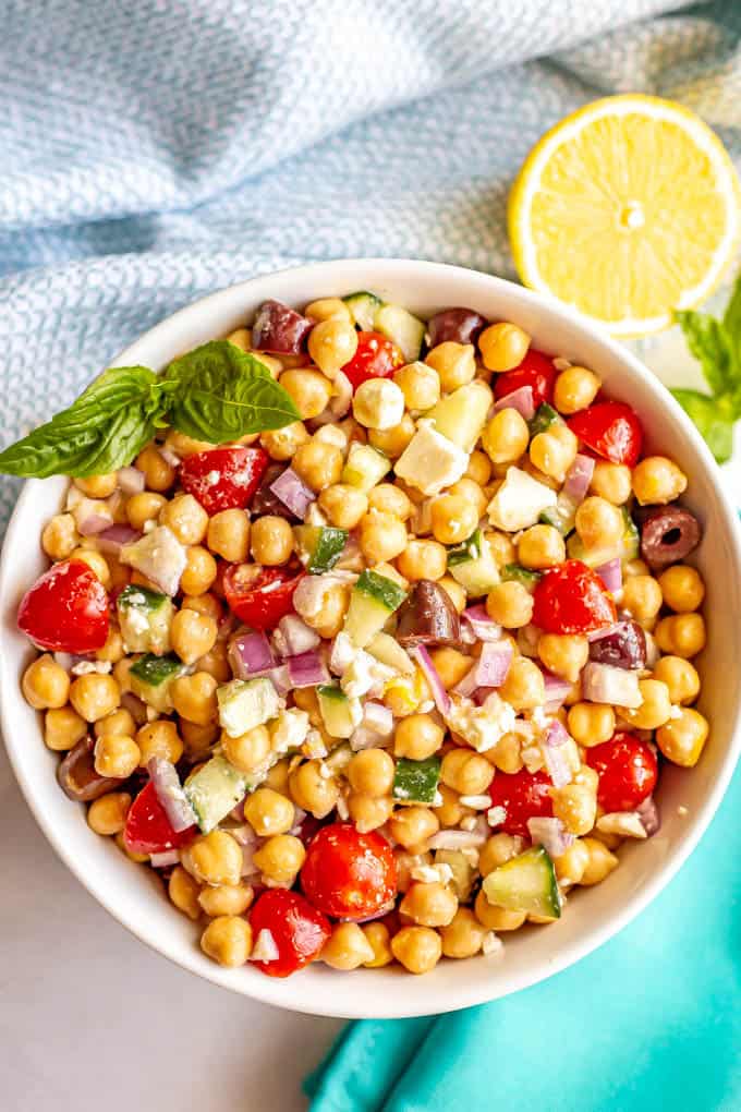 A large white serving bowl with a colorful, fresh Mediterranean style chickpea salad with a cut lemon and basil nearby