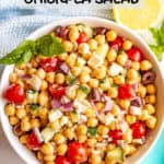 A colorful bowl of chickpea salad with tomatoes, cucumbers, olives and onions with a sprig of basil and a text box on top