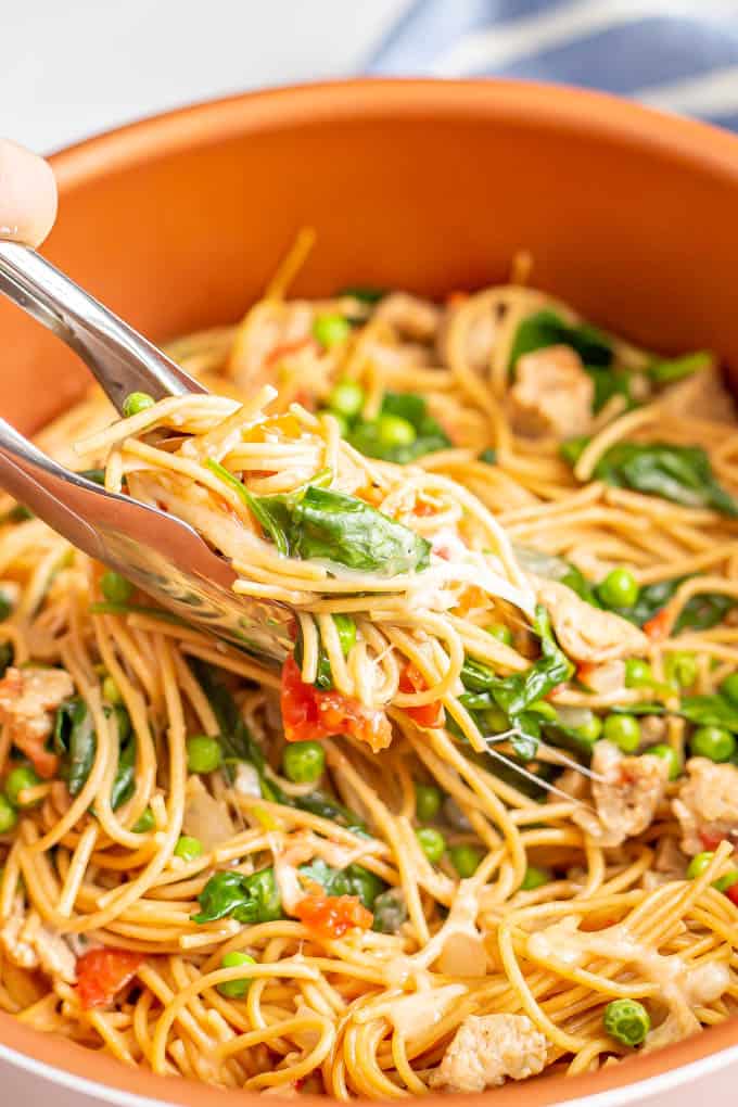 Tongs picking up a scoop of spaghetti noodles with chicken sausage, spinach and peas from a large copper pot