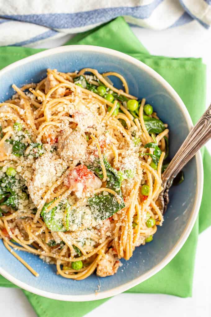 A blue and white bowl of pasta with chicken sausage, spinach and peas and Parmesan cheese on top with two forks resting in the bowl