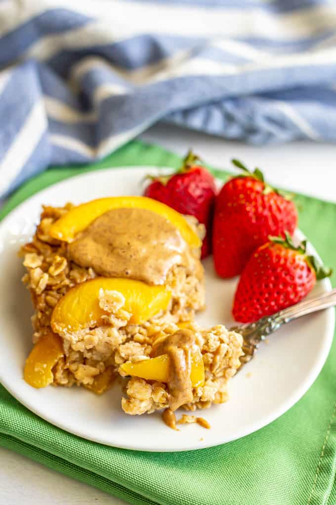 A slice of baked oatmeal with peaches and almond butter served on a plate with strawberries and a forkful being scooped up