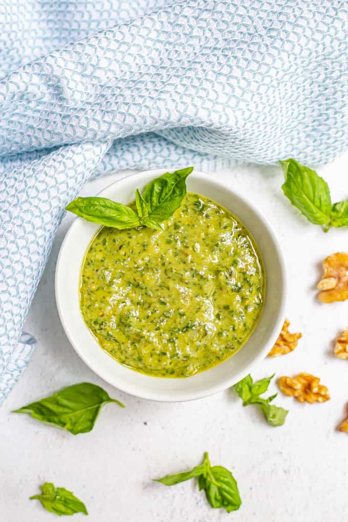 A white bowl of fresh basil pesto with sprigs of homemade pesto and walnut pieces scattered nearby