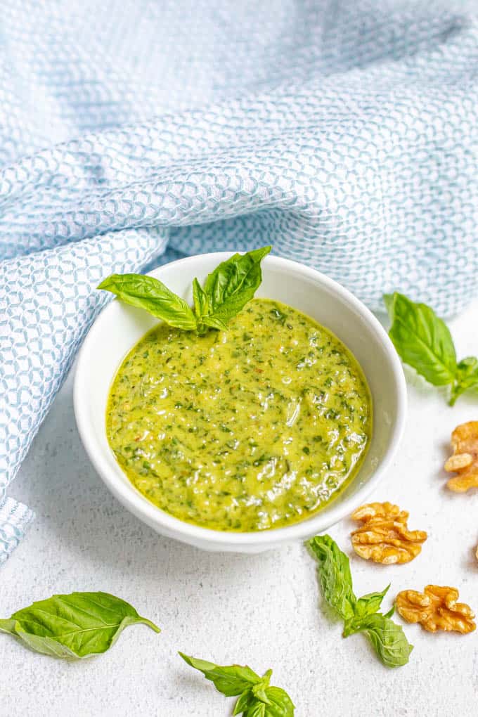 A bowl of fresh basil pesto with sprigs of fresh pesto and walnut pieces scattered nearby