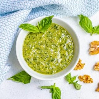 A white bowl of fresh basil pesto with sprigs of fresh pesto and walnut pieces scattered nearby