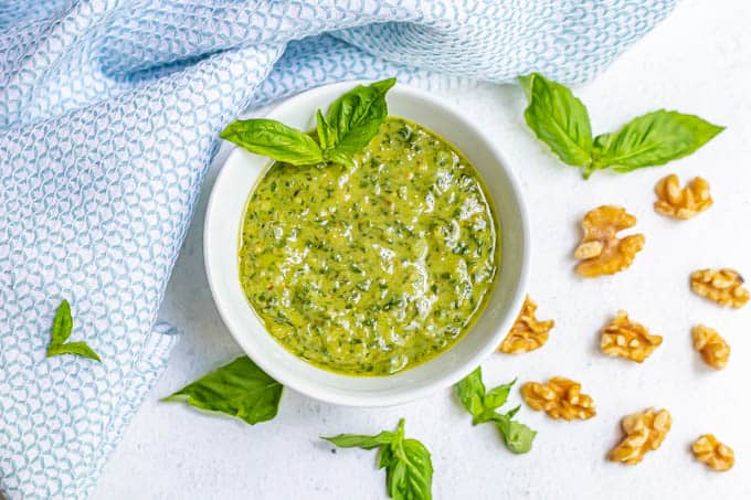 A white bowl of fresh basil pesto with sprigs of fresh pesto and walnut pieces scattered nearby