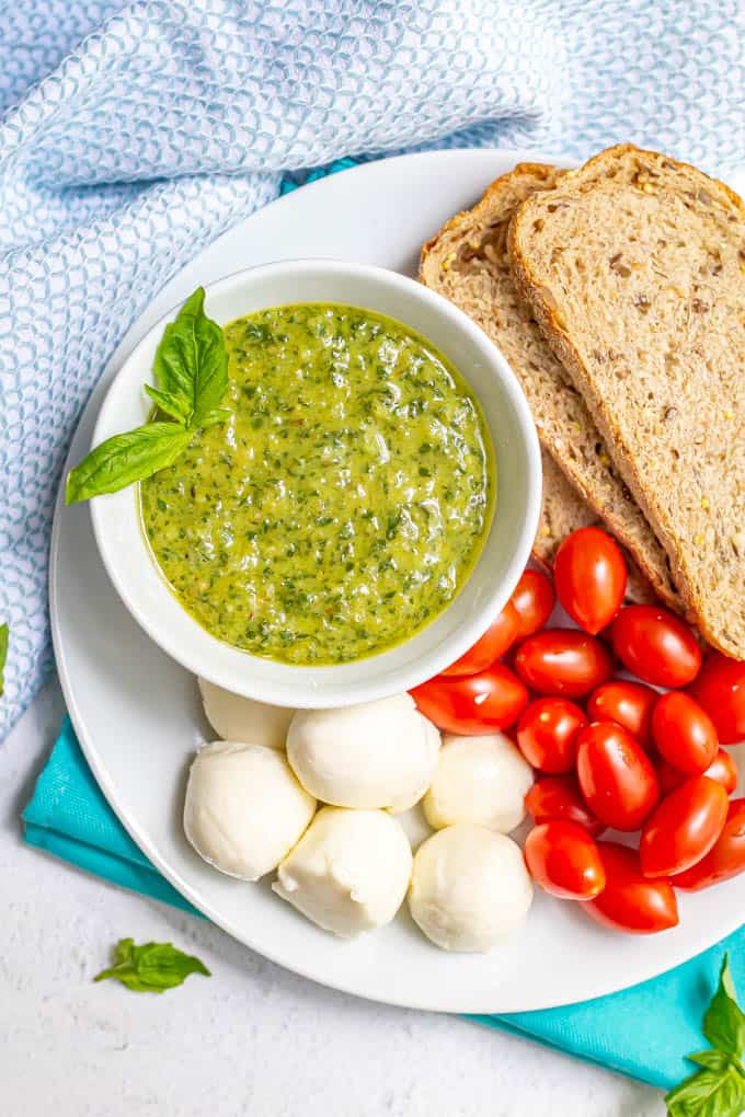 Pesto served in a white bowl on a white plate of whole grain bread, tomatoes and mozzarella balls