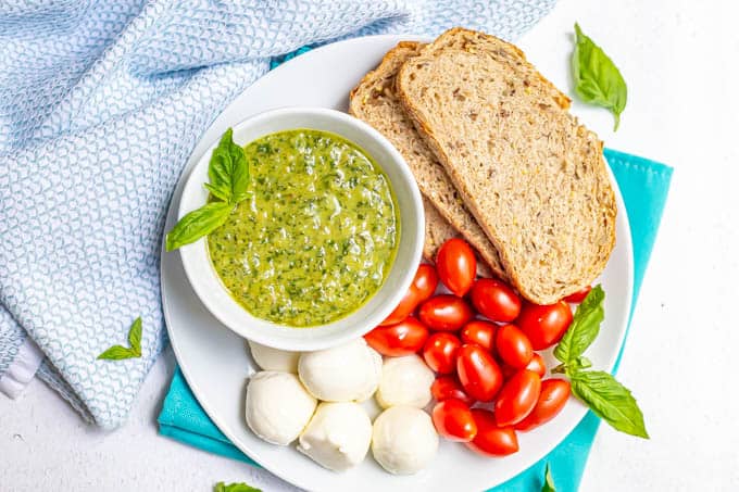 Pesto served in a white bowl on a plate of whole grain bread, tomatoes and mozzarella balls with sprigs of basil around